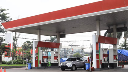 Ein Auto tankt an einer Tankstelle mit roter und weißer Beschriftung, umgeben von Bäumen und Gebäuden.