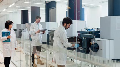 two scientists wearing white lab coats working in a laboratory facing a large rectangular analytical instrument